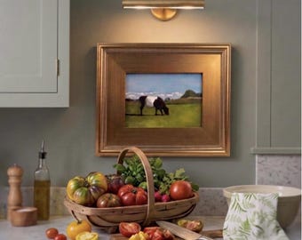 Op de afbeelding: Een houten mand gevuld met verse producten, waaronder vleestomaten, een rode tomaat en groene kruiden. Een houten snijplank met gesneden tomaten en een mes staat ervoor. Een ingelijst schilderij van een koe hangt onder een messing wandlamp.