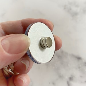May include: A white ceramic round button with a blue and white design on the front and a silver magnet on the back.