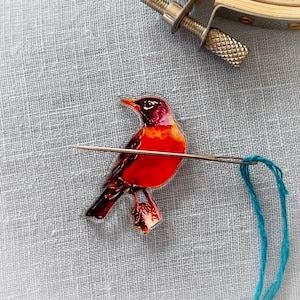 May include: A vibrant red and black bird design is the focal point of this embroidery project. A needle with blue thread is threaded through the fabric, showcasing the beginning of a detailed craft. The bird is perched on a light gray fabric, with a metal embroidery hoop in the background.