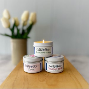 May include: Three soy wax candles in white tins with blue and white striped lids. The candles are labeled "Goûté Bleu" and have different scents: "Outdoor Shower", "Florist", and "Kitchen Table".