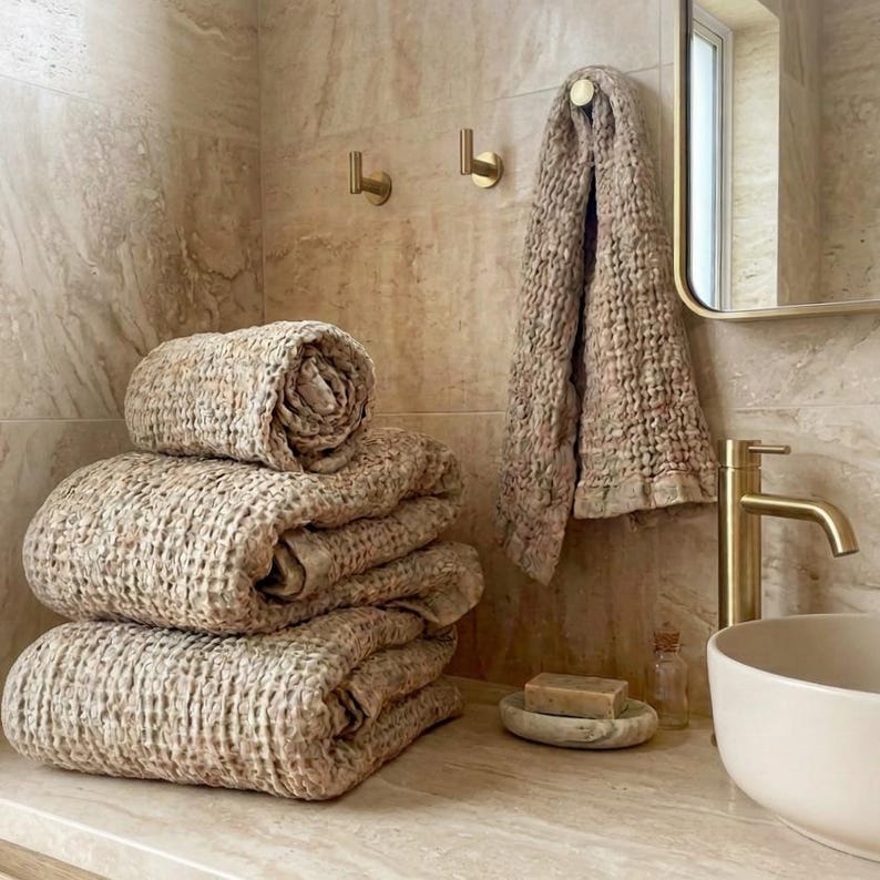 May include: A stack of three folded beige waffle-textured towels and one hanging towel, positioned next to a white ceramic vessel sink with a gold faucet, a bar of soap on a stone dish, and two gold wall hooks against a beige marble wall.