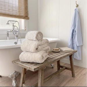 May include: A stack of three folded beige waffle-textured towels resting on a rustic wooden bench. Beside the towels sits a small bar of soap on a dish and a closed book. A light blue robe hangs on a wall hook near a white clawfoot bathtub.