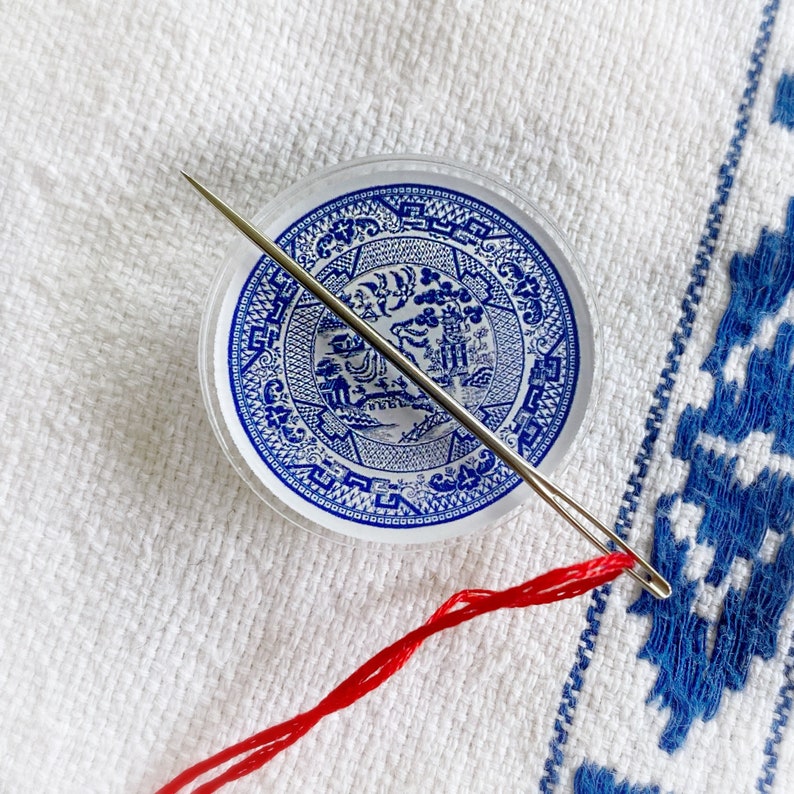 May include: A close-up of a blue and white porcelain plate with a floral design. A silver needle is threaded through the plate and a red string is attached to the needle.