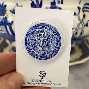 May include: A blue and white ceramic pin featuring a traditional Chinese design. The pin is round and has a glossy finish. The design includes a tree, a bird, and a fence. The pin is on a white card with the text "OwletteBleu" and social media handles.