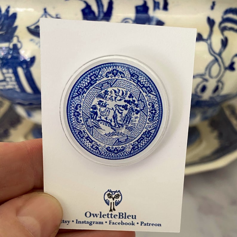 May include: A blue and white ceramic pin featuring a traditional Chinese design. The pin is round and has a glossy finish. The design includes a tree, a bird, and a fence. The pin is on a white card with the text "OwletteBleu" and social media handles.