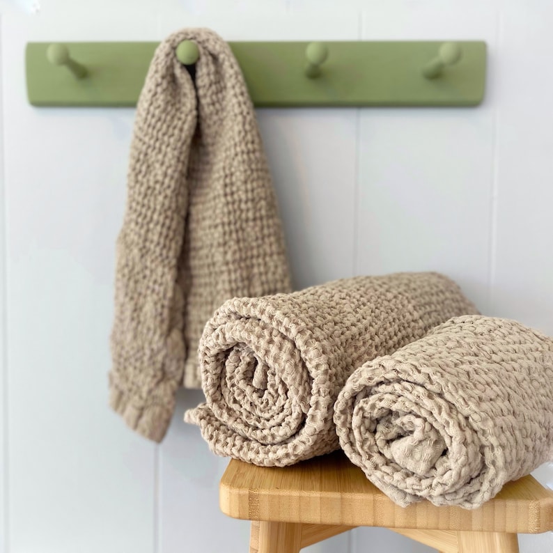 May include: Three beige waffle-weave towels. One towel hangs from a sage green wall-mounted hook rack, while two other towels are tightly rolled and stacked on top of each other. The fabric features a distinct, textured grid pattern throughout.