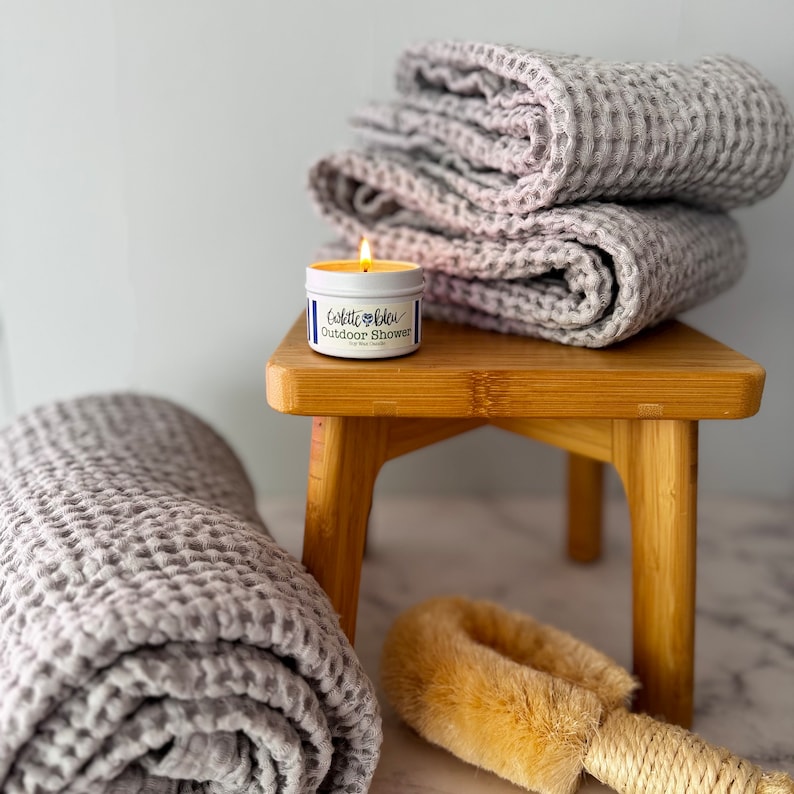 May include: A lit candle in a white tin with the label "Violette Bleu Outdoor Shower" sits on a small wooden stool. Beside it are folded grey waffle-textured towels and a long-handled natural bristle bath brush with a rope loop for hanging.