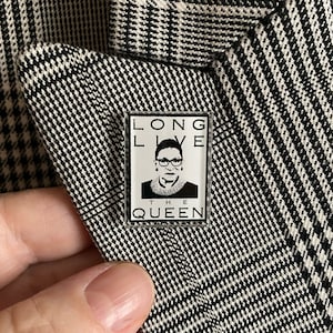 May include: A rectangular enamel pin featuring a black and white portrait of a woman wearing glasses and a collar. The pin has the text "LONG LIVE THE QUEEN" in a black font. The pin is on a black and white plaid blazer.