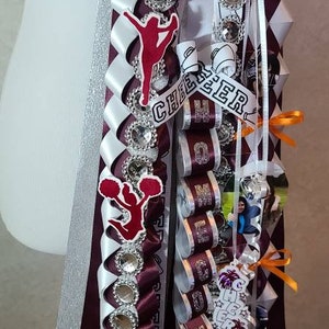 K&ouml;nnte beinhalten: Ein individuelles Homecoming-Mum mit weinroten, wei&szlig;en und silbernen B&auml;ndern. Verzierungen umfassen Strassakzente, Cheerleader-Figuren und das Wort "CHEER". Das Mum hat eine gro&szlig;e Blume und dekorative B&auml;nder.