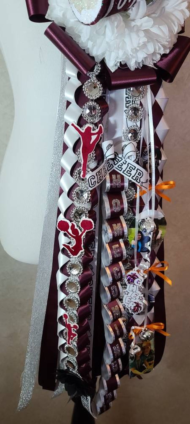 K&ouml;nnte beinhalten: Ein individuelles Homecoming-Mum mit weinroten, wei&szlig;en und silbernen B&auml;ndern. Verzierungen umfassen Strassakzente, Cheerleader-Figuren und das Wort "CHEER". Das Mum hat eine gro&szlig;e Blume und dekorative B&auml;nder.