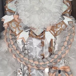 K&ouml;nnte beinhalten: Eine dekorative Mum, mit einer gro&szlig;en wei&szlig;en Blume in der Mitte, umgeben von Schichten aus silbernen, pfirsichfarbenen und goldglitzernden B&auml;ndern. Str&auml;nge aus Perlen und Schleifen erg&auml;nzen das festliche Design.