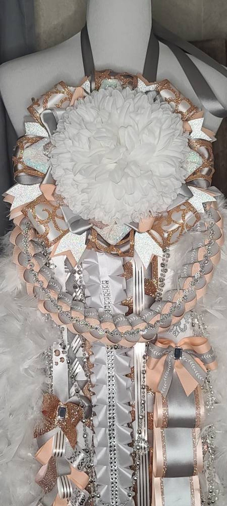 K&ouml;nnte beinhalten: Eine dekorative Mum, mit einer gro&szlig;en wei&szlig;en Blume in der Mitte, umgeben von Schichten aus silbernen, pfirsichfarbenen und goldglitzernden B&auml;ndern. Str&auml;nge aus Perlen und Schleifen erg&auml;nzen das festliche Design.