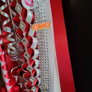 K&ouml;nnte beinhalten: Dekoratives rotes und silbernes Band mit einer Strasskette, einem Knopf mit rosa Lippen und dem Wort "OMG". Das Band ist mit roten Federn und einem wei&szlig;en Band mit rot-wei&szlig;en Rautenmustern verziert.