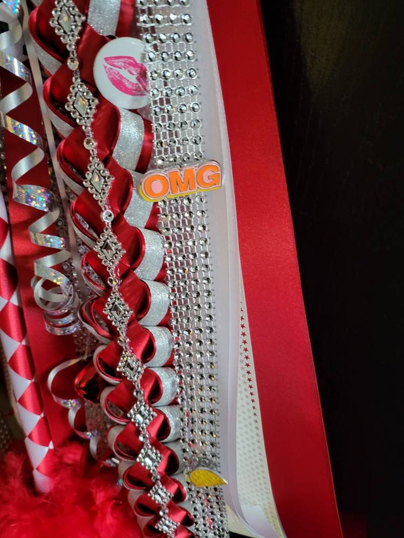 K&ouml;nnte beinhalten: Dekoratives rotes und silbernes Band mit einer Strasskette, einem Knopf mit rosa Lippen und dem Wort "OMG". Das Band ist mit roten Federn und einem wei&szlig;en Band mit rot-wei&szlig;en Rautenmustern verziert.