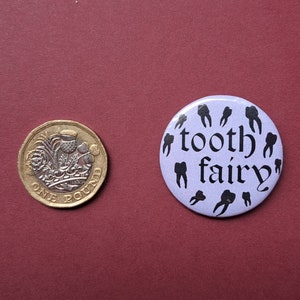 Pu&ograve; includere: Una moneta da una sterlina accanto a un bottone lilla con le parole "tooth fairy" e illustrazioni di denti neri. La moneta &egrave; dorata e argentata con un design dettagliato.