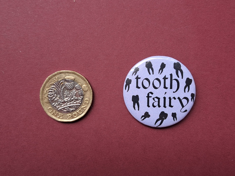 Pu&ograve; includere: Una moneta da una sterlina accanto a un bottone lilla con le parole "tooth fairy" e illustrazioni di denti neri. La moneta &egrave; dorata e argentata con un design dettagliato.