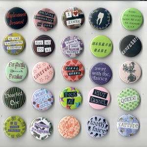May include: A selection of circular pin badges with diverse designs and text. The badges display phrases such as "Unpleasant Dreams!", "I Zines", "Horror Babe", and "Queer". Colours include black, pink, green, and purple. Some badges feature illustrations.