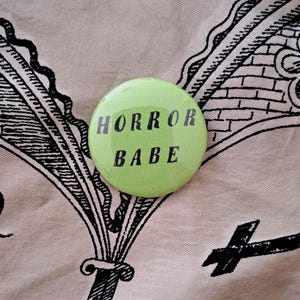 Pu&ograve; includere: Un bottone rotondo verde chiaro con la scritta nera "HORROR BABE". Il bottone &egrave; posto su un tessuto rosa chiaro con illustrazioni nere.