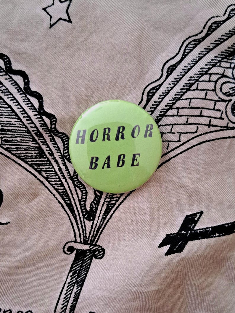Pu&ograve; includere: Un bottone rotondo verde chiaro con la scritta nera "HORROR BABE". Il bottone &egrave; posto su un tessuto rosa chiaro con illustrazioni nere.