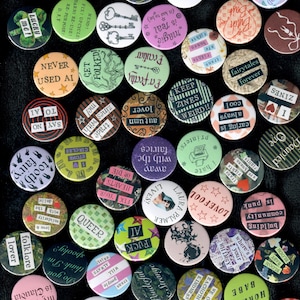 Pu&ograve; includere: Una collezione di spille rotonde con vari disegni e testi. Le spille presentano frasi come "Lucky Me!", "Queer" e "Fairytales Forever". I colori includono rosa, verde e nero.