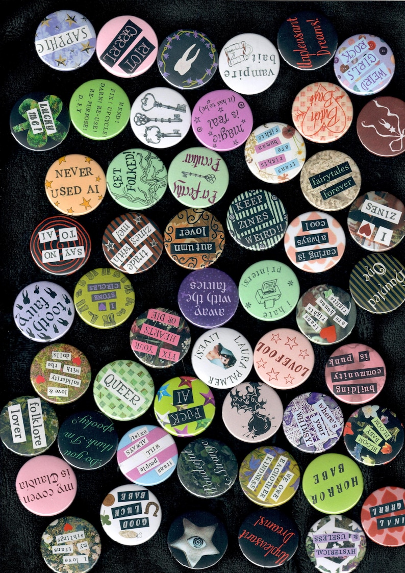 Pu&ograve; includere: Una collezione di spille rotonde con vari disegni e testi. Le spille presentano frasi come "Lucky Me!", "Queer" e "Fairytales Forever". I colori includono rosa, verde e nero.