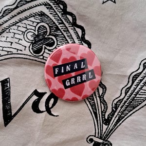 Pu&ograve; includere: Un bottone rotondo rosa con un motivo di cuori rossi e le parole "FINAL GRRRL" in lettere nere. Il bottone &egrave; posto su un tessuto color crema con motivi decorativi e testo neri.