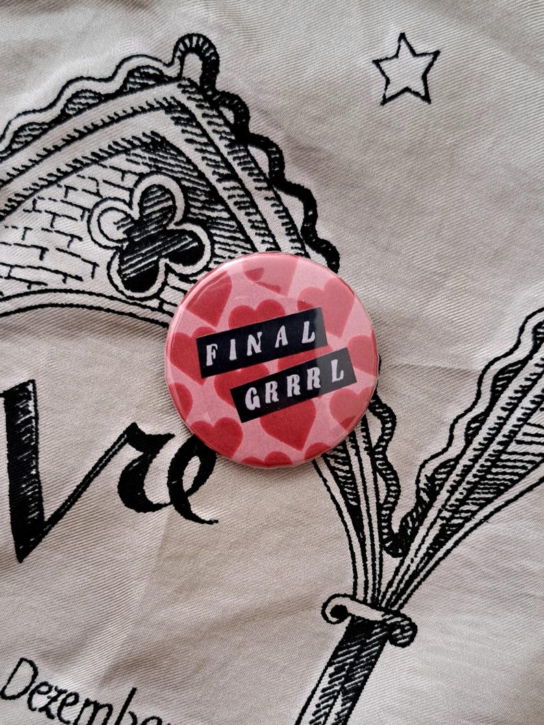 Pu&ograve; includere: Un bottone rotondo rosa con un motivo di cuori rossi e le parole "FINAL GRRRL" in lettere nere. Il bottone &egrave; posto su un tessuto color crema con motivi decorativi e testo neri.