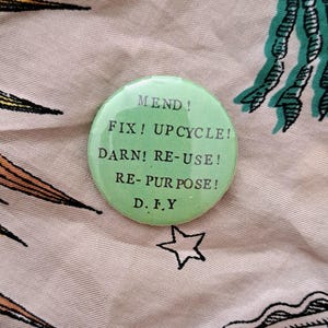 Pu&ograve; includere: Un bottone rotondo verde chiaro con le parole "MEND! FIX! UPCYCLE! DARN! RE-USE! RE-PURPOSE! D.I.Y" in testo nero. Il bottone &egrave; su un tessuto fantasia con una stella e altri disegni.
