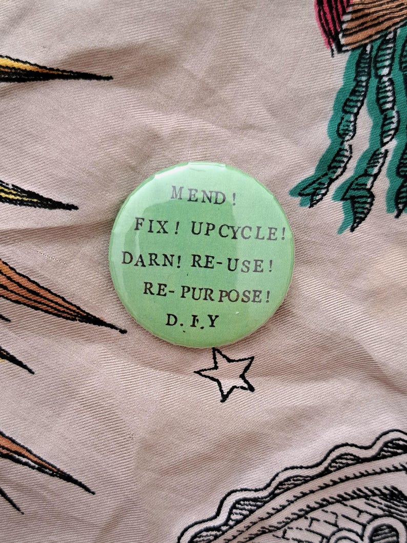 Pu&ograve; includere: Un bottone rotondo verde chiaro con le parole "MEND! FIX! UPCYCLE! DARN! RE-USE! RE-PURPOSE! D.I.Y" in testo nero. Il bottone &egrave; su un tessuto fantasia con una stella e altri disegni.