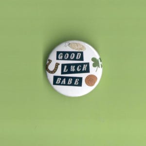 May include: A white button with the words "GOOD LUCK BABE" in black, featuring a horseshoe, shamrock, and coin illustrations. The button is set against a green backdrop.