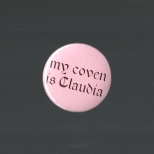 May include: A circular, pale pink button featuring the phrase "my coven is Claudia" in dark brown gothic script. The button is set against a dark grey backdrop. A simple, yet striking statement piece.