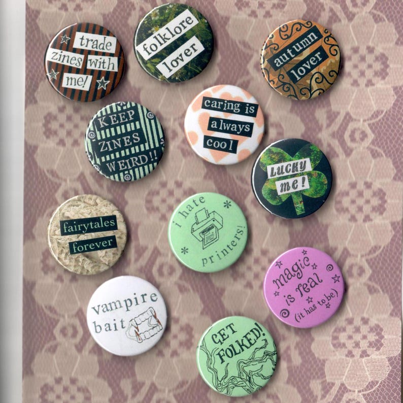 Pu&ograve; includere: Una collezione di bottoni rotondi e colorati con vari testi e disegni grafici. I bottoni presentano frasi come "trade zines with me!", "folklore lover" e "magic is real". Lo sfondo &egrave; un tessuto a motivi simili a pizzo.