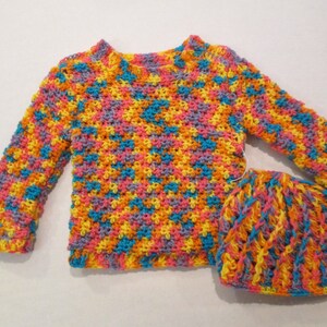 Puede incluir: Un conjunto de punto de ganchillo colorido en tonos de rosa, amarillo, azul y naranja. El suéter tiene mangas largas y cuello redondo. El gorro es de estilo beanie.