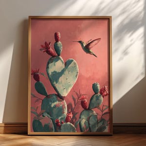 May include: A framed art print featuring a heart-shaped cactus with a hummingbird in flight. The artwork has a pink and teal color palette, with red flowers and a wooden frame. The print is leaning against a wall.