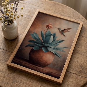 Puede incluir: Una impresi&oacute;n art&iacute;stica enmarcada que presenta una pintura a la acuarela de una planta de agave verde azulado en una maceta marr&oacute;n, un colibr&iacute; y una flor. La obra de arte tiene una paleta de colores c&aacute;lidos y est&aacute; sobre un fondo marr&oacute;n. El marco es de madera clara.