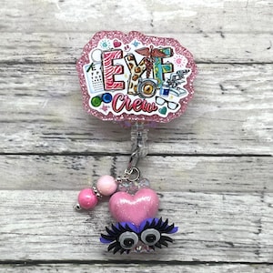 May include: A pink glitter badge reel with the words "EYE Crew" and eye-related graphics. The reel has a pink heart charm with eyelashes and a beaded accent.