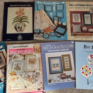 May include: A collection of cross stitch patterns featuring various themes, including floral, baby, and seasonal designs. The patterns are printed on colorful paper with titles like "Orchid in Imari Vase", "Just For Baby", and "Four Season Sampler".