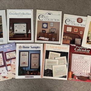 Può includere: Una collezione di nove schemi di ricamo a punto croce con vari temi, tra cui un alfabeto da cucina, un sampler per la casa, un posto nel tuo cuore, un calendario perpetuo e una collezione di grilli. Gli schemi sono stampati su carta bianca con illustrazioni e testo colorati.