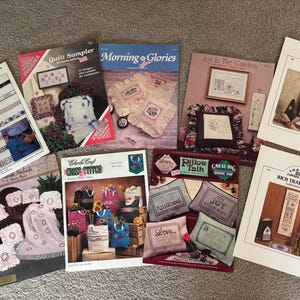 May include: A collection of 10 cross-stitch patterns featuring various designs, including a quilt sampler, floral motifs, and inspirational quotes. The patterns are printed on white paper with colourful illustrations and text.
