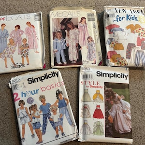 May include: Five sewing pattern envelopes from McCall's, New Look, and Simplicity brands. The envelopes feature illustrations and photographs of various clothing items including robes, pajamas, dresses, and children's outfits with printed text.