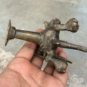 Pode incluir: Puxador de porta antigo em bronze com um design escultural detalhado. O puxador apresenta uma base circular, um corpo cilíndrico e uma figura ornamental com forma de animal. O bronze tem uma pátina desgastada, sugerindo idade. Uma peça única para decoração vintage.