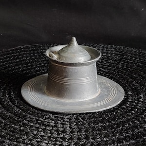 May include: A silver metal inkwell with a pointed lid, sitting on a black circular woven mat.