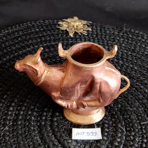 May include: A copper-coloured metal vase shaped like a cow with a round opening at the top. The vase is sitting on a black woven mat. The label on the mat reads "MT 599".