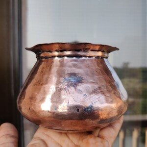 May include: A small, copper pot with a rounded body and a flared rim. The pot has a hammered finish and shows signs of age and use.