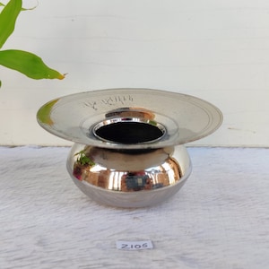 Puede incluir: Un cuenco de metal plateado con un borde ancho y plano y una base redonda y bulbosa. El cuenco tiene una pequeña abertura en el centro del borde. El cuenco está sobre una superficie de madera blanca.