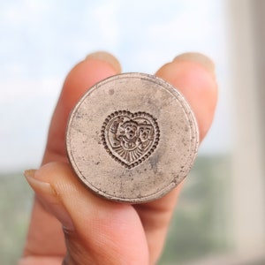 Puede incluir: Un sello de metal plateado con un diseño en forma de corazón que presenta un patrón floral detallado y tres caras dentro del corazón.