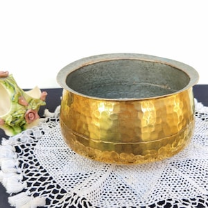 May include: A hammered gold metal bowl with a round shape and a textured surface. The bowl is sitting on a white crocheted doily with fringe.