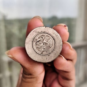 Puede incluir: Un sello de metal plateado con un diseño detallado de dos pájaros que se miran. El sello es circular y tiene un borde elevado.