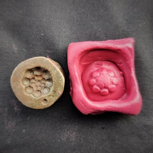 Puede incluir: Un sello de latón antiguo con un diseño circular de pequeños círculos y una impresión de cera rosa. El sello está deslustrado con un tono verde en las áreas empotradas. La impresión de cera es cuadrada y muestra el diseño en relieve.