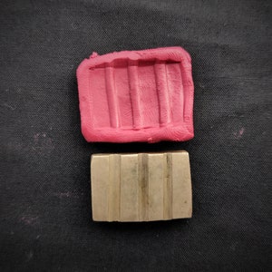 Puede incluir: Un objeto rectangular de latón con ranuras verticales se encuentra debajo de un molde rectangular rosa con ranuras a juego. Los objetos están sobre una superficie negra. El objeto de latón parece ser una hebilla o un componente.